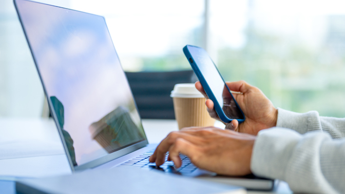 Hands holding a phone while working on a computer in sunny office